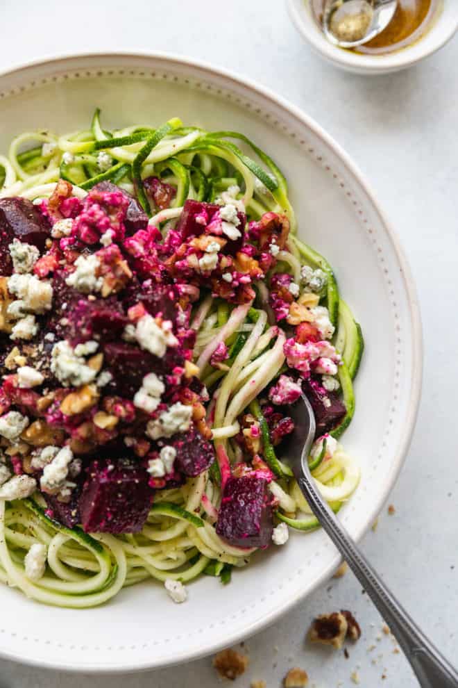 This is an overhead view of a bowl of zoodles, beets, and crumbled blue cheese. The bowl sit son a white counter and a fork is in the bowl, twirling the zoodles. 