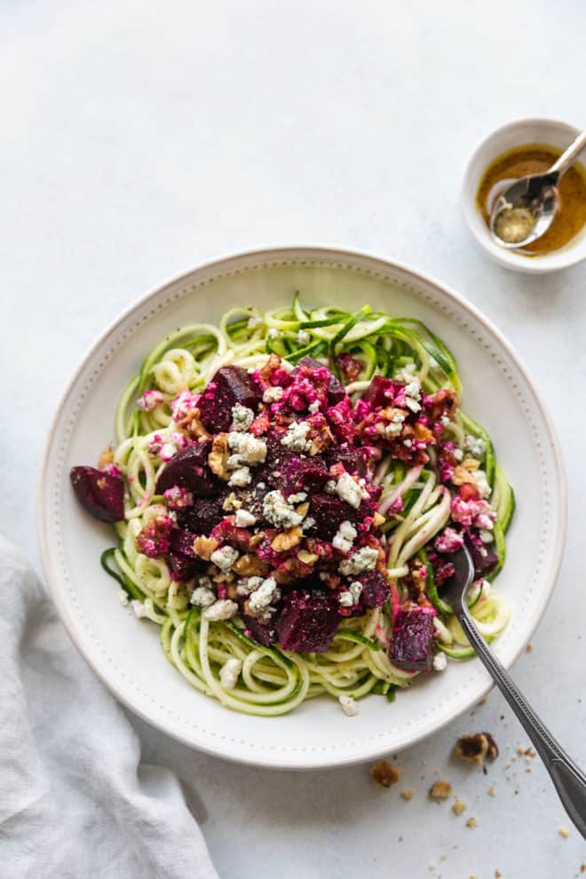 This is an overhead image of a bowl of zoodles topped with beets, blue cheese and walnuts. A fork is twirling the zoodles and leaving against the side of the bowl. 