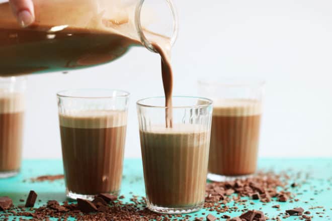 Glass pitcher filled with creamy chocolate flax milk is pouring chocolate milk into four small glass cups. The cups are on a green counter with chocolate and flax seeds scattered around them. 