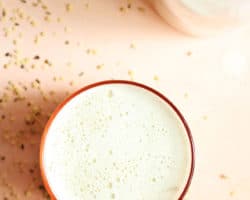 This is an overhead image of a pink glass filled with hemp milk. The glass sits on a light pink surface with hemp hearts scattered around it.