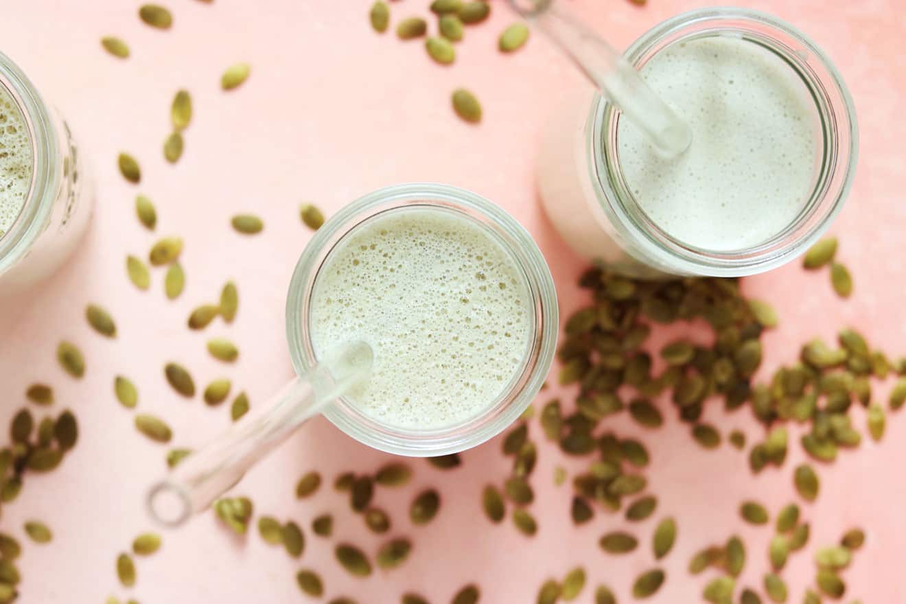 This is an overhead image of two glasses with a slightly green liquid in it. The glasses are on a light pink surface with raw pumpkin seeds scattered around. Each glass has a glass straw sticking out.