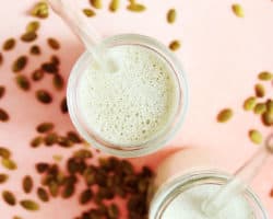 This is an overhead image of two glasses with a slightly green liquid in it. The glasses are on a light pink surface with raw pumpkin seeds scattered around. Each glass has a glass straw sticking out.