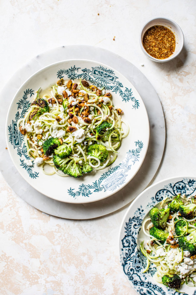 This is an overhead image of a zoodle salad with broccoli, pistachios, and crumbled cheese. A small bowl of mustard vinaigrette is off to the top left of the image. 