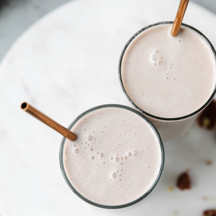 This is an overhead image of two glasses of pecan nut milk. The glasses sit on a marble circle cutting board on a grey surface. There are copper straws in the cups of pecan milk. Raw pecans are around the glasses of milk.