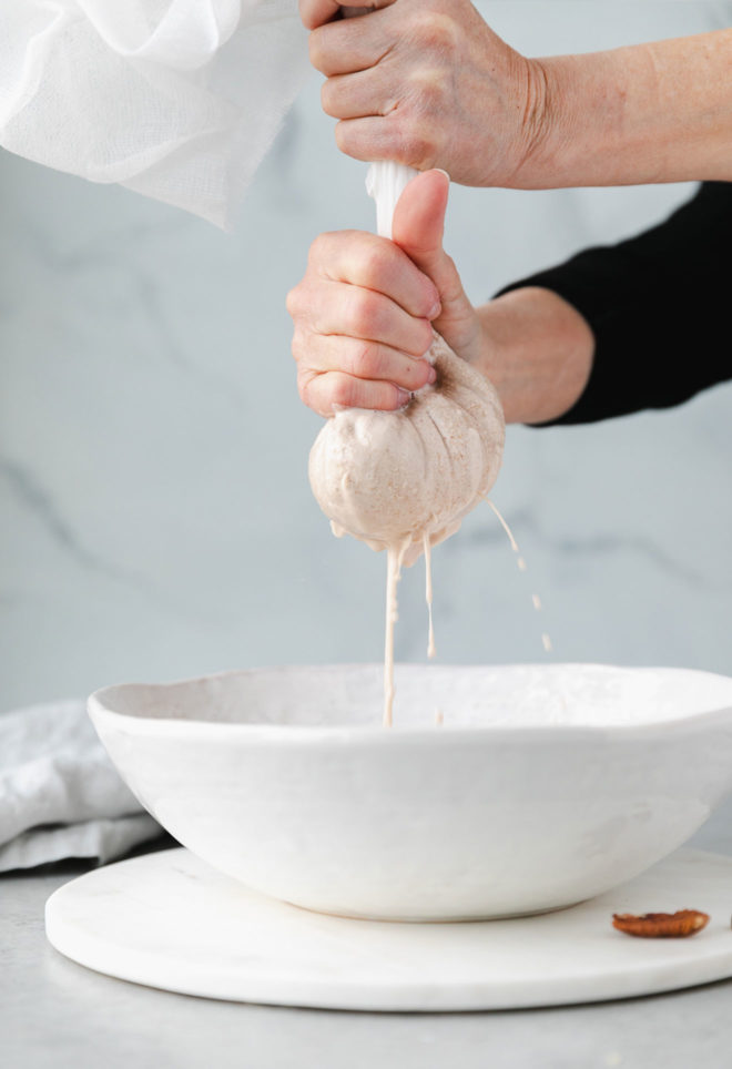 This is a side view of hands squeezing liquid out of a nut milk bag into a white bowl. The bowl is on a white charger on a marble counter. The background is marble and there is a grey tea towel off the the right. One pecan sits next to the bowl. 