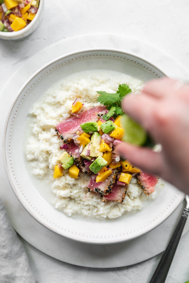 This is an overhead image of a bowl filled with coconut rice. On top of the rice is ahi tuna, mango chunks, avocado chunks, and fresh cilantro. A hand is blurred in the image squeezing lime juice on top of the meal. More mango salsa is in the top left corner of the image. The bowl sits on a marble charger on a white counter.