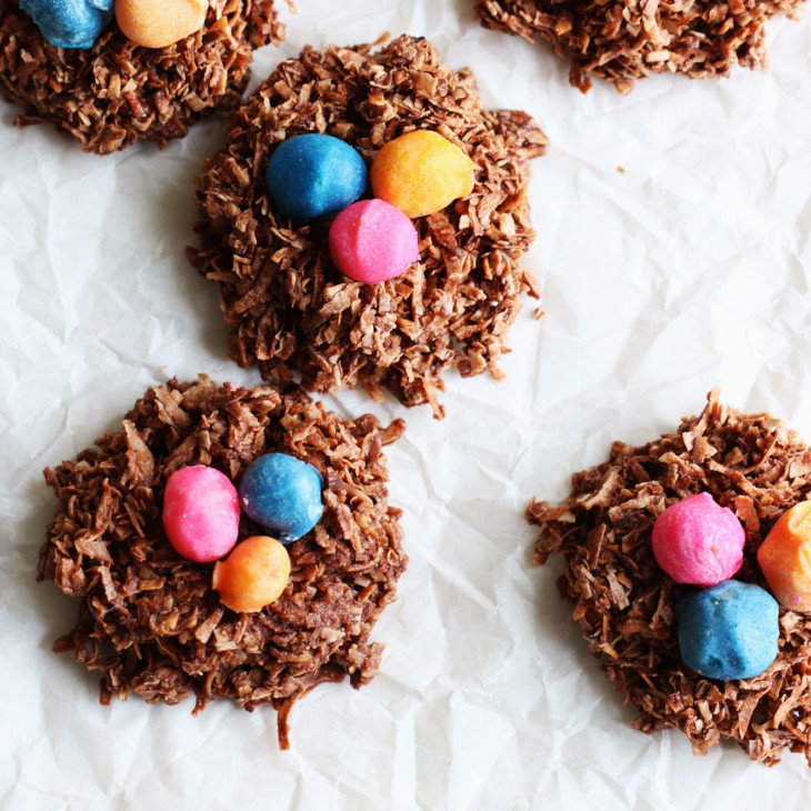 This is an overhead image of chocolate coconut circle cookies with colorful nuts in the center of each one. The cookies resemble a bird's nest and are sitting on a white piece of parchment paper.