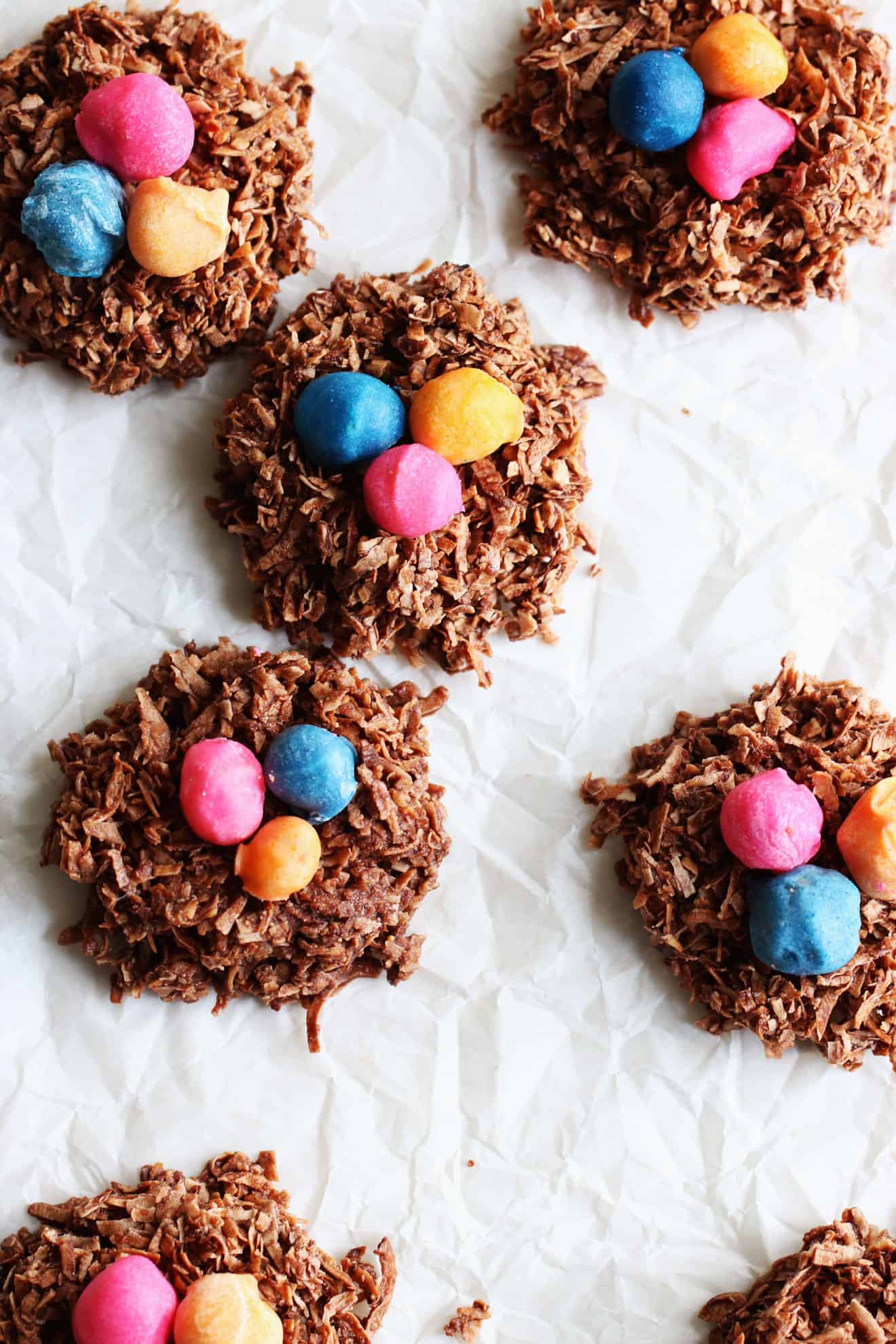 This is an overhead image of chocolate coconut birds nest cookies with colorful nuts in the center of each one. The cookies resemble a bird's nest and are sitting on a white piece of parchment paper.