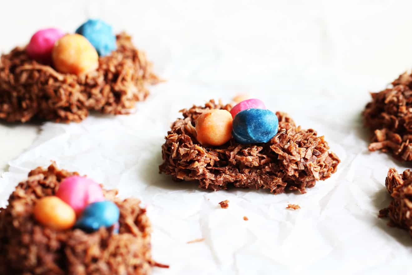 This is a side view of chocolate coconut bird's nest cookies with colorful nuts in the center of each one. The cookies resemble a bird's nest and are sitting on a white piece of parchment paper. A bite is taken out of the cookie and more cookies are blurred to the side and background.
