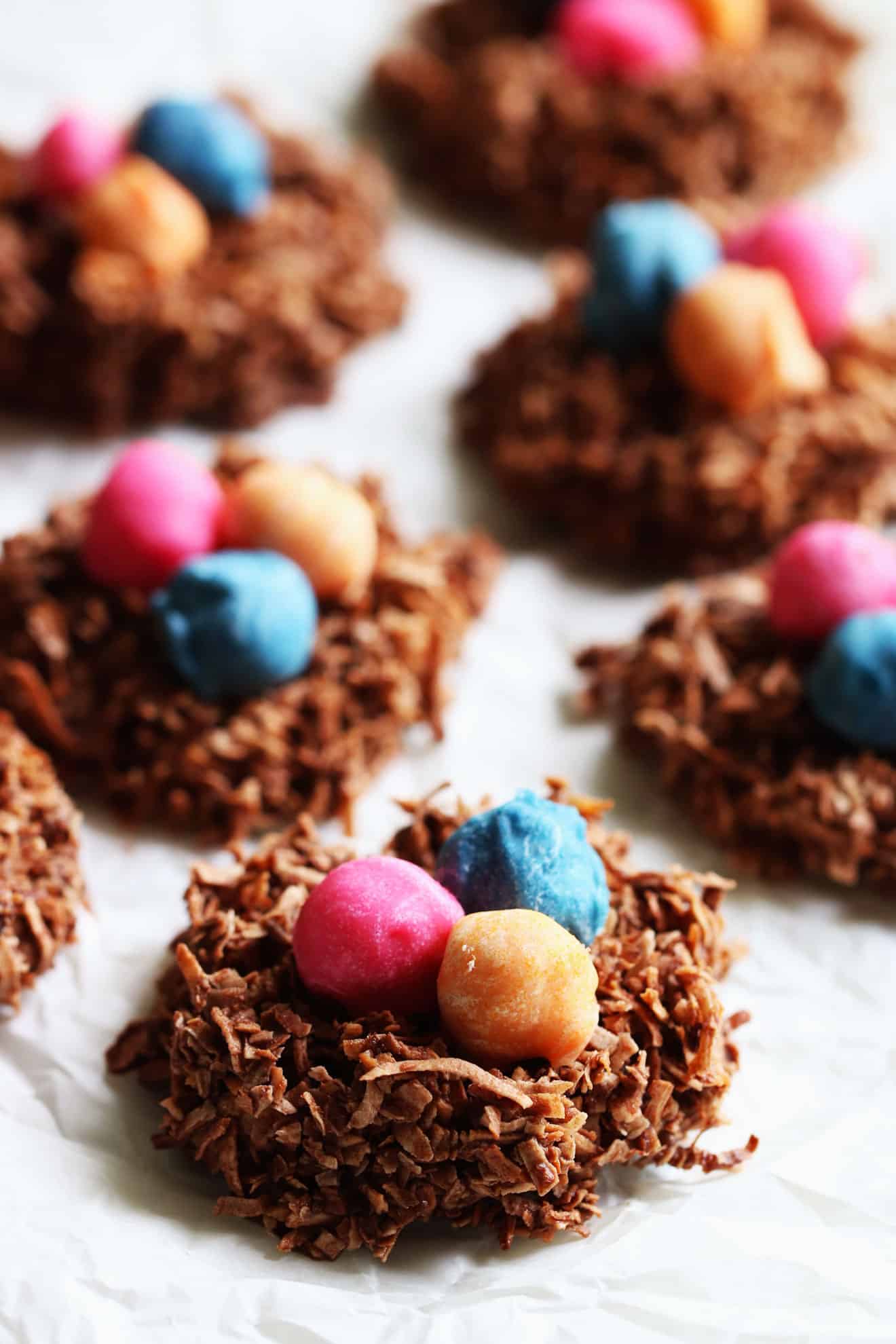 This is a side view of chocolate coconut birds nest cookies with colorful nuts in the center of each one. The cookies resemble a bird's nest and are sitting on a white piece of parchment paper. More cookies are blurred in the background.