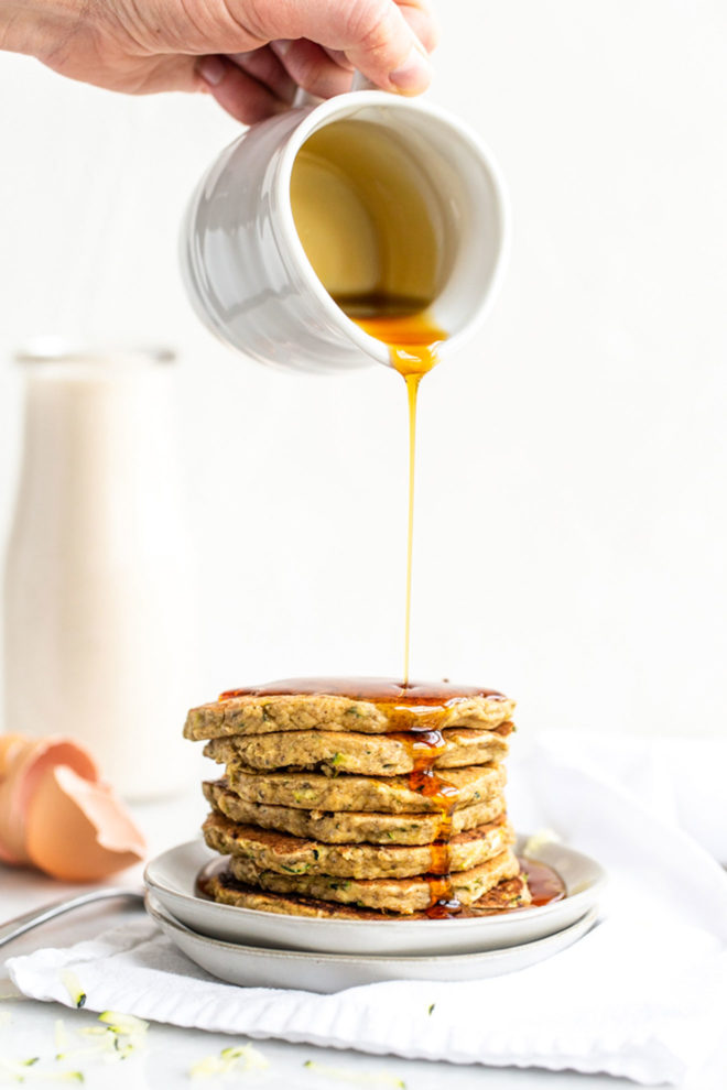 A hand is pouring syrup on top of a pile of zucchini pancakes on a plate. Milk and egg shells are blurred in the background. You can see specs of green in the pancakes. 