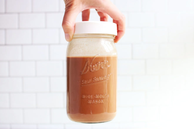 This is a side view of a hand holding a mason jar. The hand is coming in from the top of the image holding onto the lid. Inside the mason jar is a chocolate milk. White subway tile is in the background. 