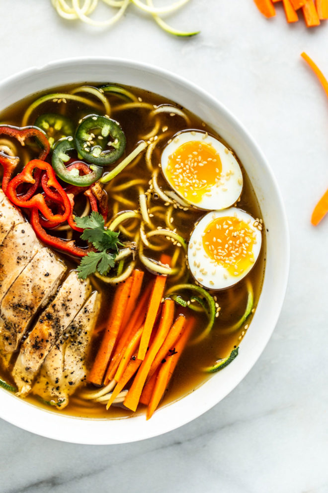 This is an overhead image of a bowl of ramen. The bowl has broth, zoodles, veggies, and a soft boiled egg. The bowl sits on a white marble counter with more veggies around it.