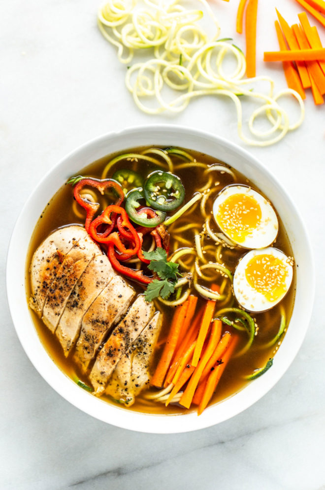 This is an overhead image of ramen soup with peppers, chicken, and an egg. More veggies are on the counter to the top right of the image. 