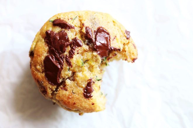 This is an overhead image of a chocolate chunk zucchini muffin with a bite taken out. The muffin sits on a white counter. 
