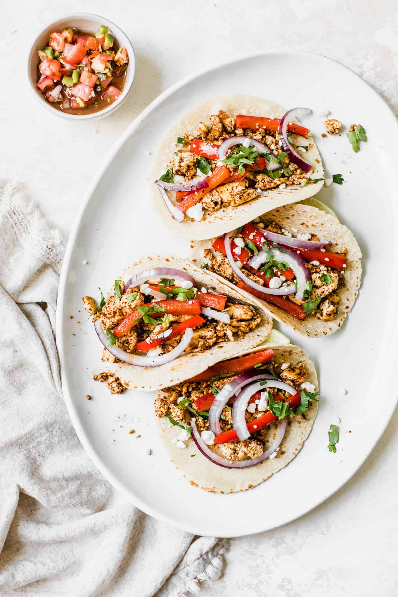 This is an overhead image of a white oval plate with four tacos on it. The tacos are loaded with crumbled tofu and slices of bell pepper, red onion pieces, crumbled goat cheese and cilantro. The plate sits on a white surface with a small bowl of chunky salsa to the top left of the image and a beige tea towel on the lefthand side of the image.