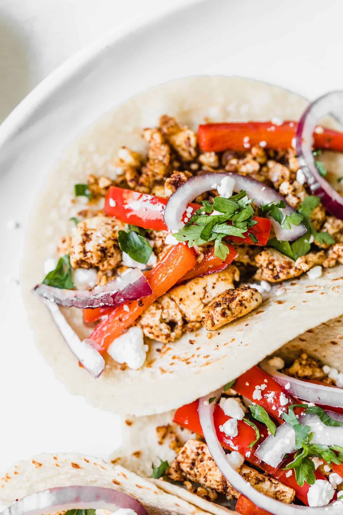 This is a closeup of tacos with crumbled spiced tofu, topped with red bell pepper strips and red onion. The tacos have crumbled cheese with cilantro on top. The taco is nestled next to another taco and on a white plate.