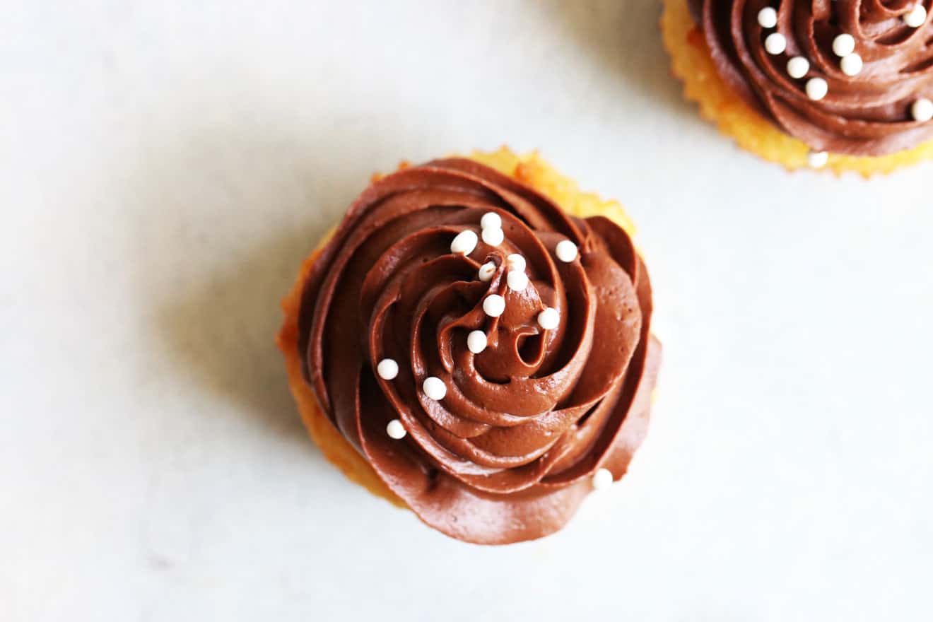 This is an overhead image of a vanilla cupcake with chocolate frosting and white ball sprinkles. The cupcake sits on a white surface with another cupcake in the upper right-hand corner of the image.