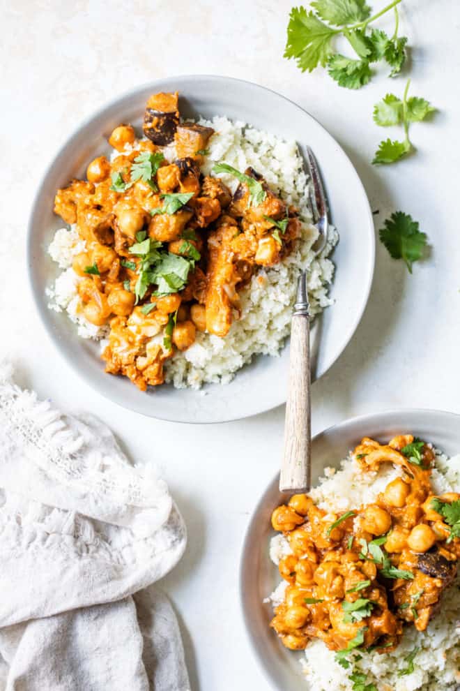This is an overhead image of two bowl filled with tikka masala on top of garlic cauliflower rice. One bowl has a fork in it. The second bowl is in the bottom right corner of the image, The bowls sit on a white counter. Fresh cilantro is in the top right of the image and some chopped cilantro leaves are sprinkled on top of each dish. A white tea towel is in the bottom left corner of the image. 