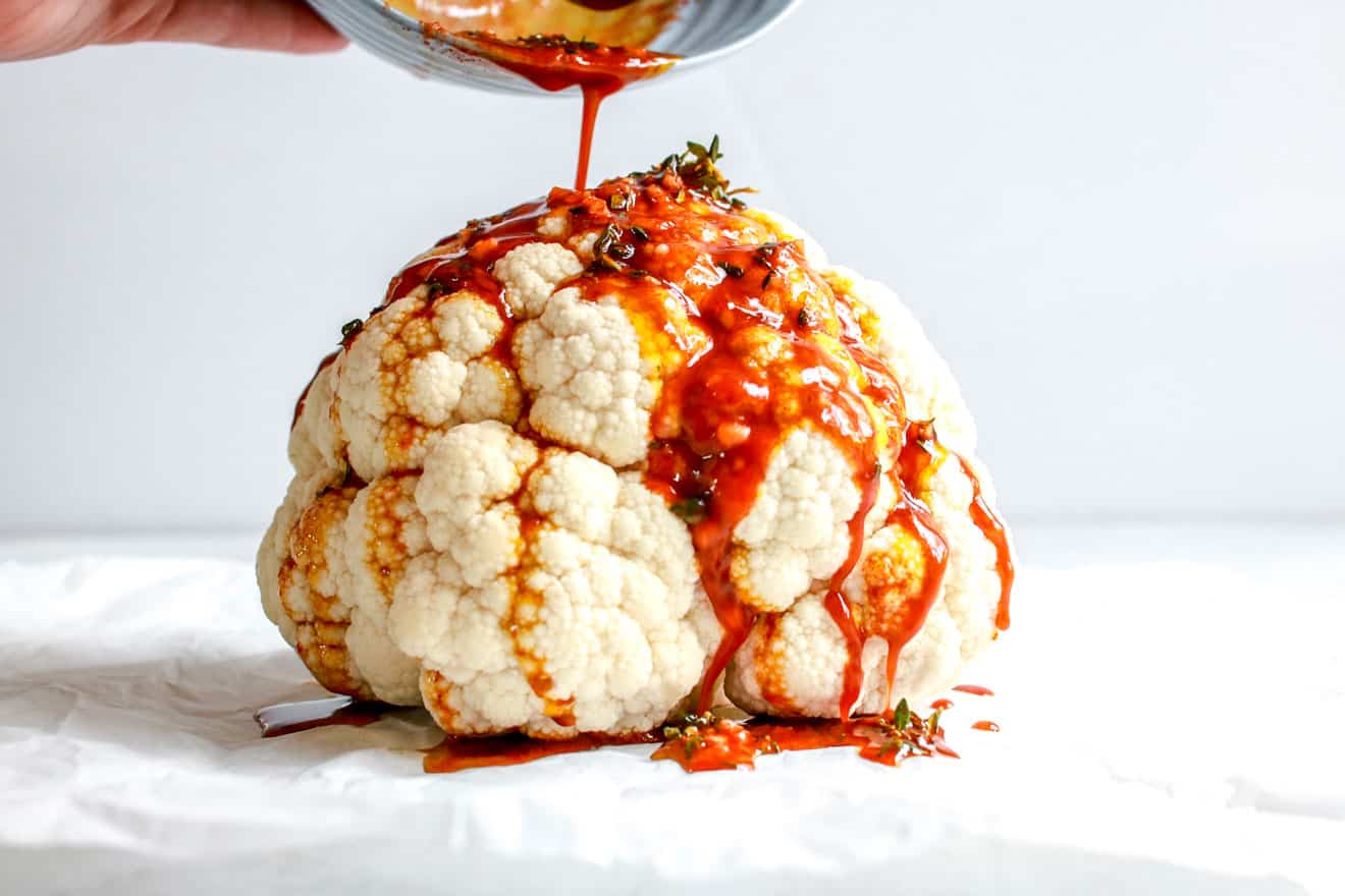 This image is a side view of a whole head of cauliflower sitting on a white counter with a white background. A hand is holding a bowl that is pouring the marinade on top of the cauliflower The marinade drizzles down the cauliflower head onto the counter.