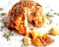 This image is a side view of a whole head of cauliflower with a drizzle of sauce on top, thyme leaves, sesame seeds, and a wedge sliced from the whole head. The head of cauliflower sides on a white piece of parchment paper on a white counter.
