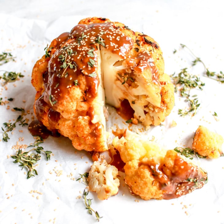 This image is a side view of a whole head of cauliflower with a drizzle of sauce on top, thyme leaves, sesame seeds, and a wedge sliced from the whole head. The head of cauliflower sides on a white piece of parchment paper on a white counter.