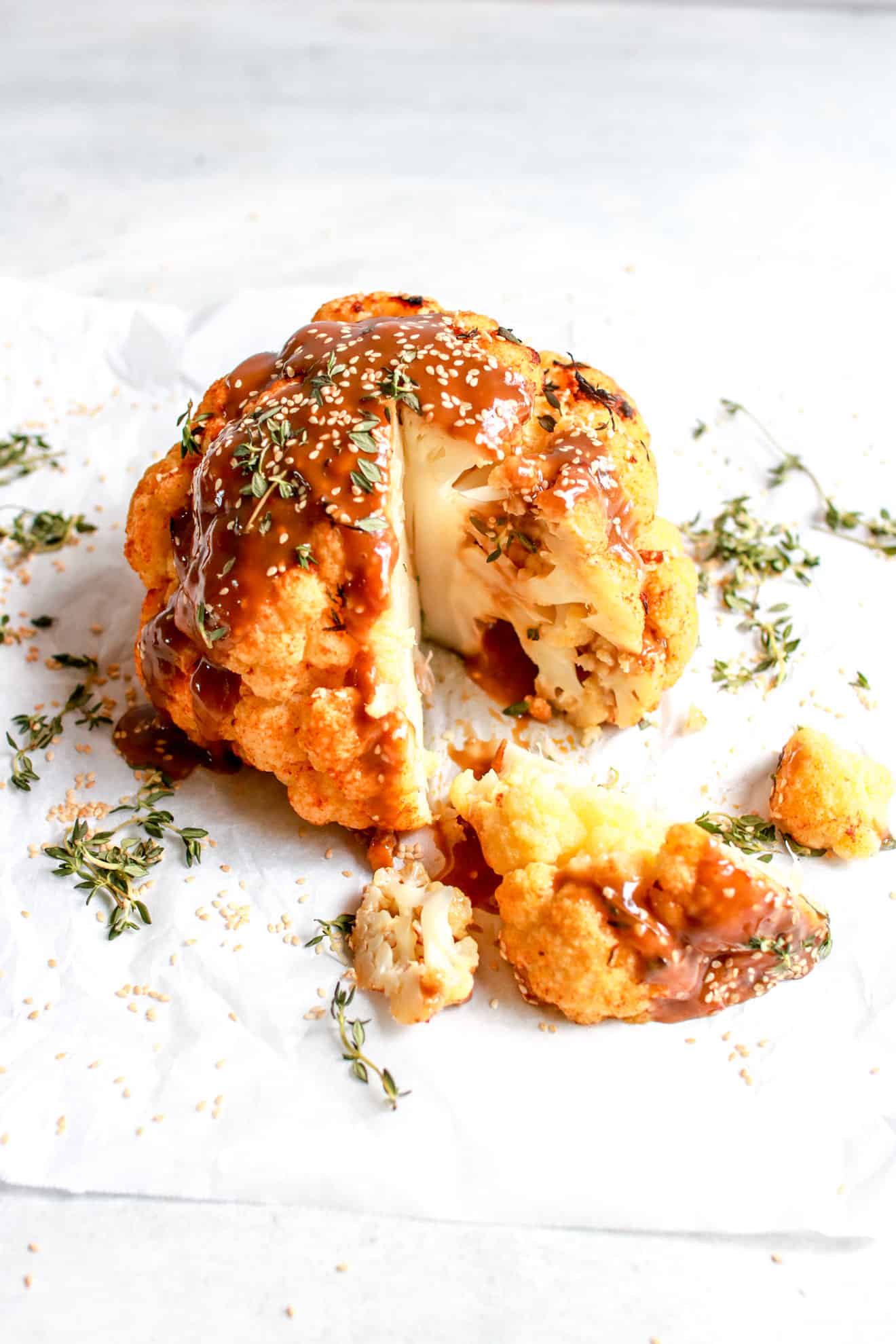 This image is a side view of a whole head of cauliflower with a drizzle of sauce on top, thyme leaves, sesame seeds, and a wedge sliced from the whole head. The head of cauliflower sides on a white piece of parchment paper on a white counter. 