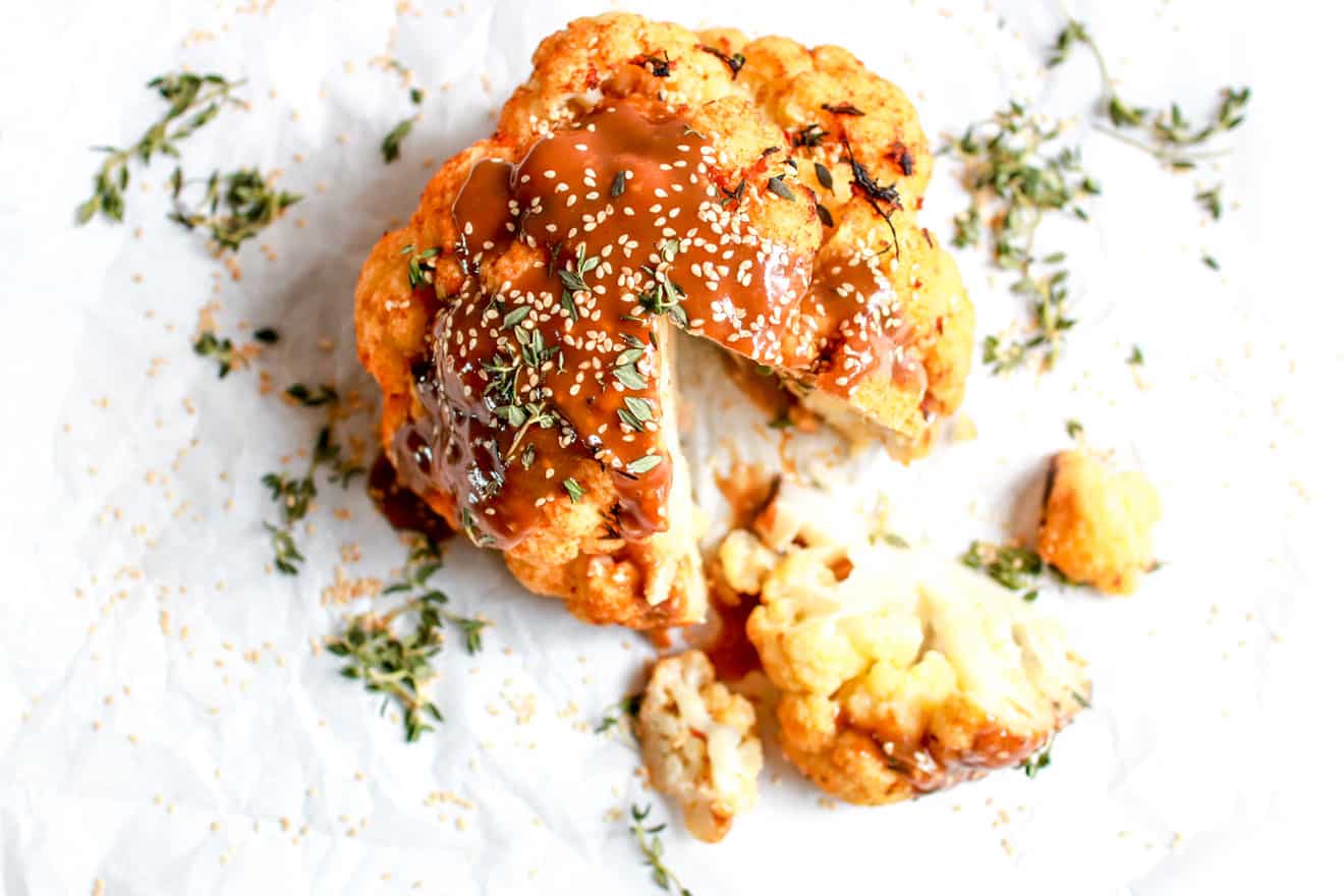 This is an overhead image of a whole head of cauliflower with a wedge cut out. The cauliflower sits on a white counter and is drizzled with a sauce, thyme leaves, and sesame seeds. 