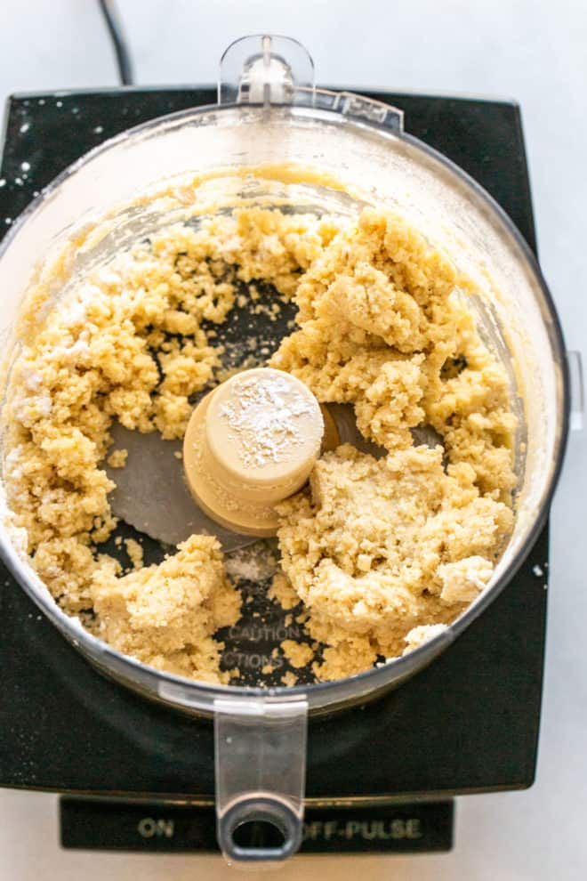 This is an overhead image of crumbly raw crust in a food processor. The food processor has a black base and sits on a white counter.