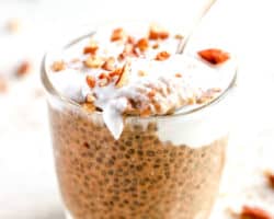 small glass cup with pumpkin chia pudding, topped with coconut cream, chopped pecans, and a spoon scooping out some, resting on the top of the glass