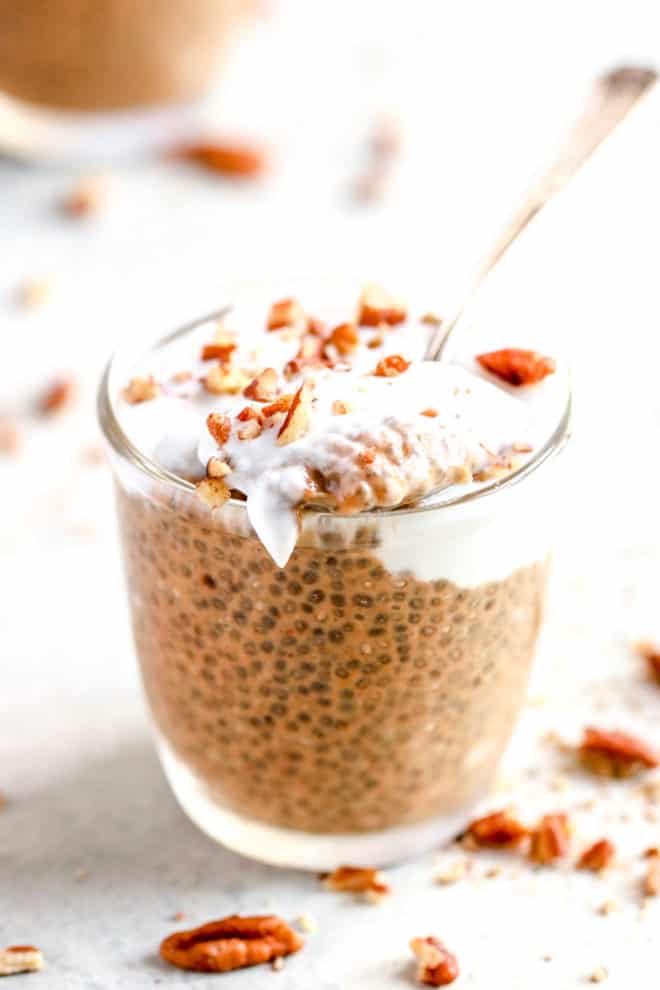 small glass cup with pumpkin chia pudding, topped with coconut cream, chopped pecans, and a spoon scooping out some, resting on the top of the glass