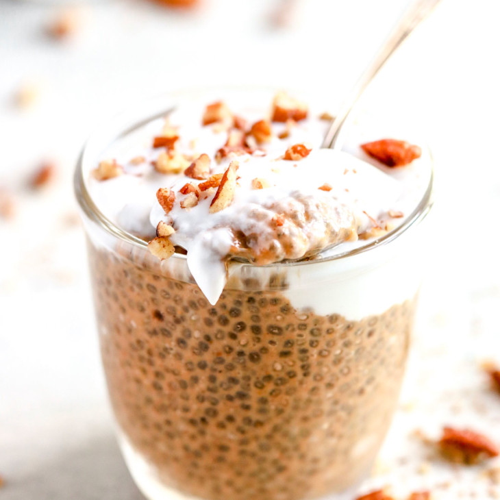 small glass cup with pumpkin chia pudding, topped with coconut cream, chopped pecans, and a spoon scooping out some, resting on the top of the glass
