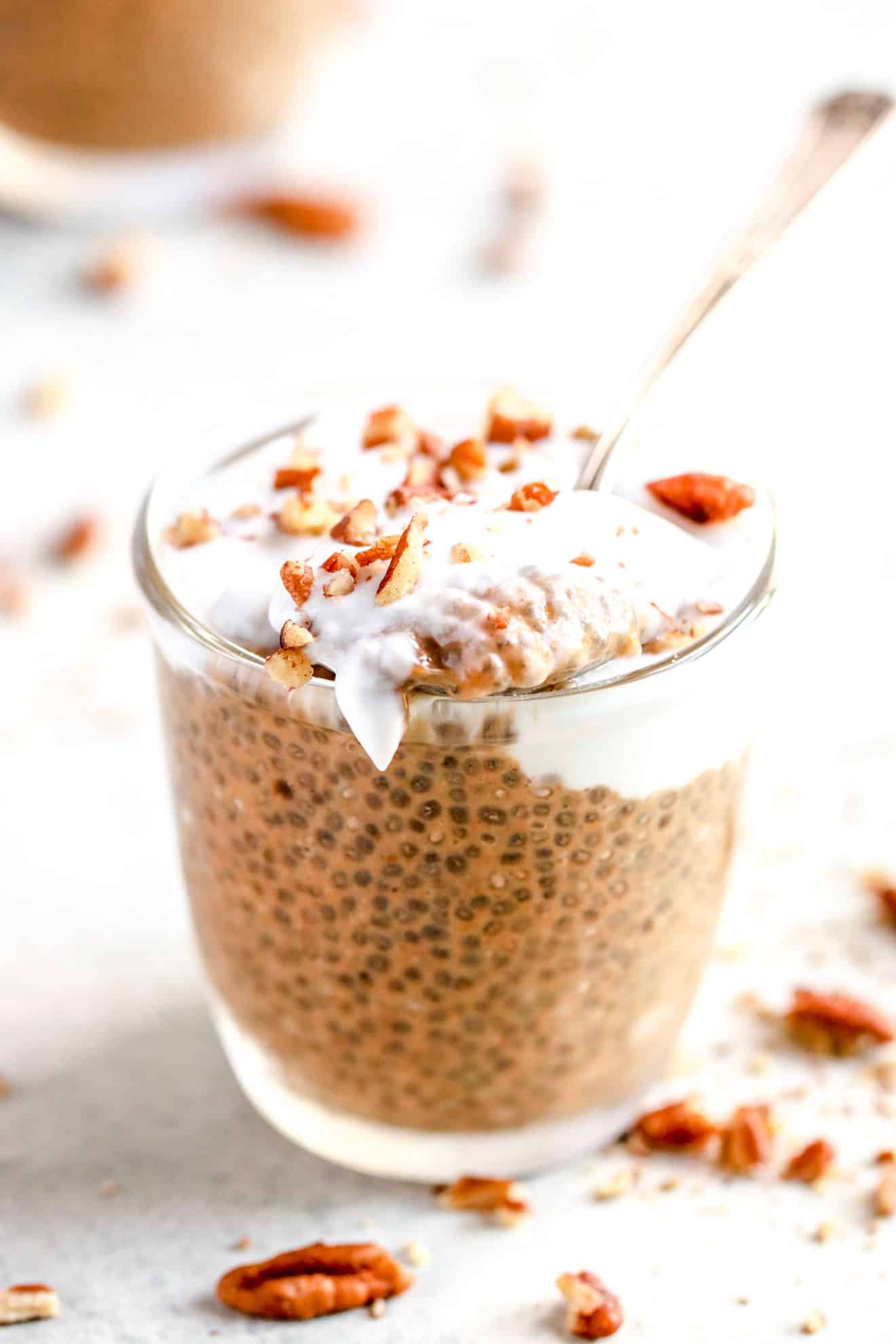 small glass cup with chia pudding, coconut cream, chopped pecans, and a spoon taking a scoop out