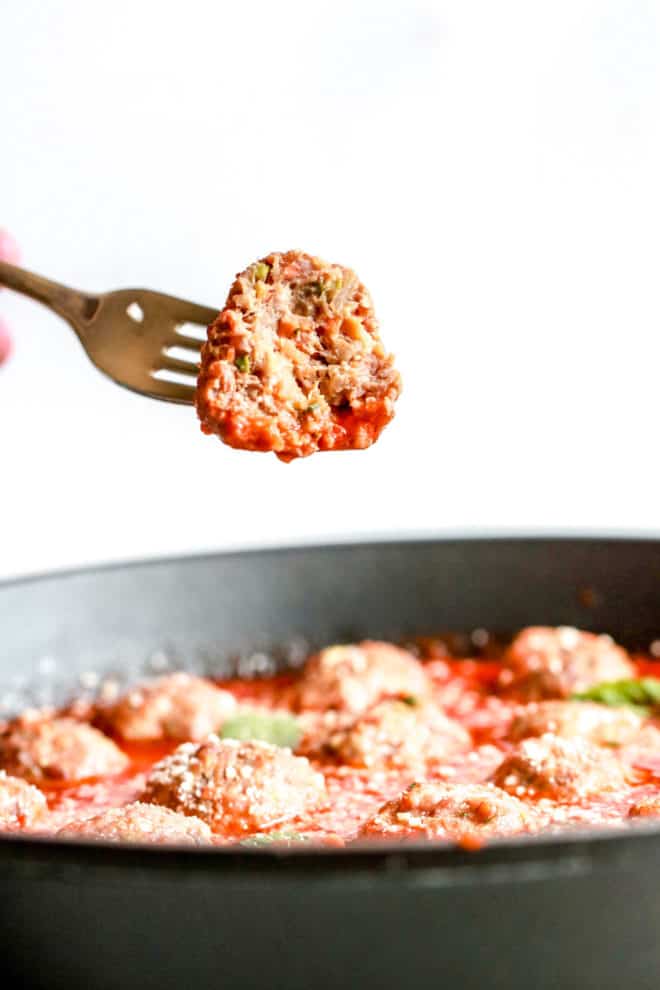 A fork is holding up a meatball with a bite taken out of it. The meatball is being held above a pan full of other meatballs. 