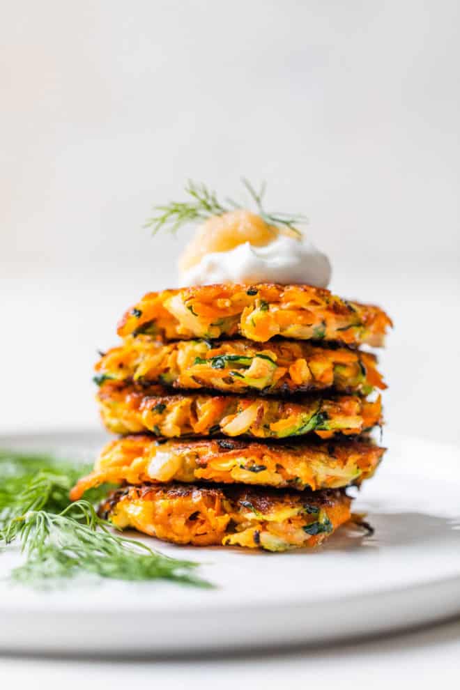 This is a stack of carrot and zucchini fritters on a white counter with a white background. Fresh dill is to the left of the stack. The fritter stack is topped with sour cream and applesauce. 