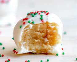 side view of a sugar cookie truffle coated in chocolate and red and green sprinkles with a bite taken out of the truffle. the truffle sites on a white counter with more sprinkles scattered around it.