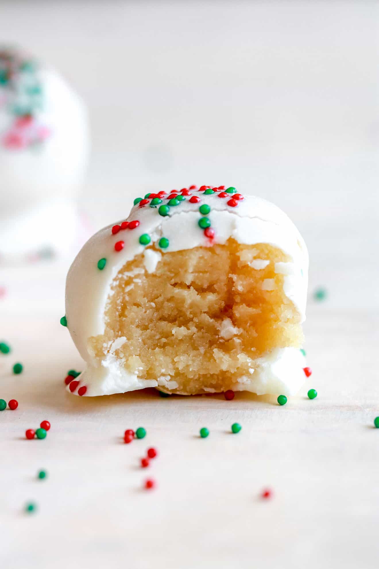 side view of a sugar cookie truffle coating with white chocolate and sprinkles with a bite taken out of the truffle on a white counter with christmas sprinkles around it