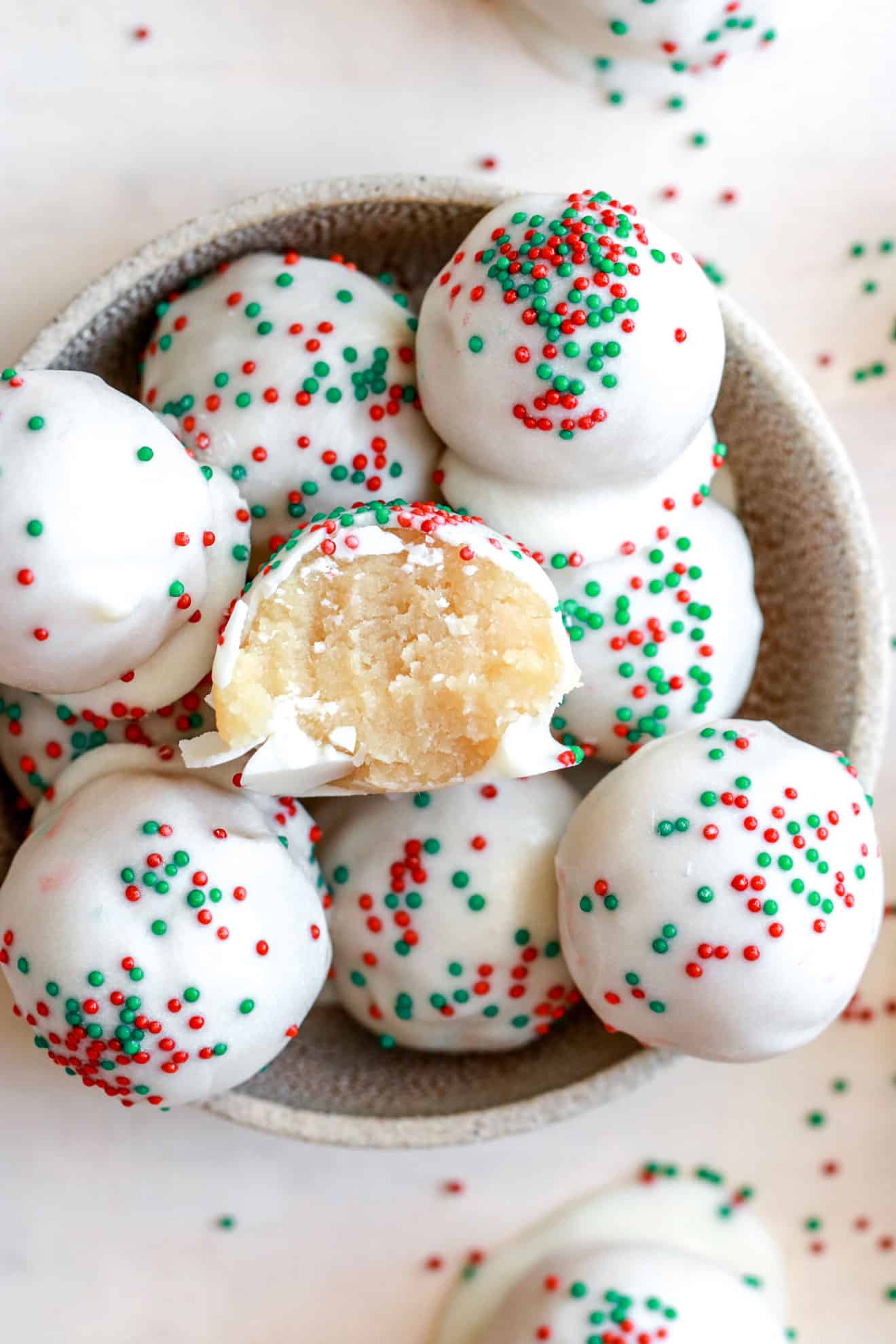 overhead view of a bowl filled with sugar cookie truffles. one truffle has a bite taken out of it. the bowl sites on a white counter with sprinkles scattered.