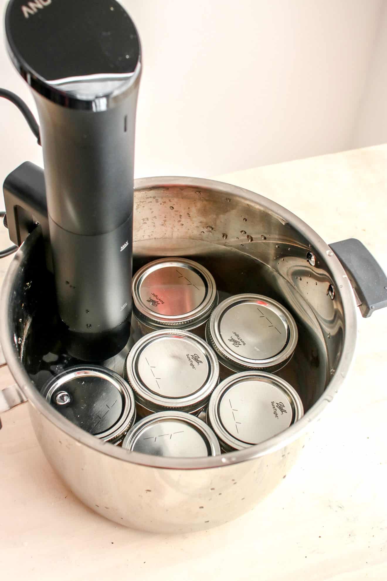 This is an overhead image o a pot filled with water and small ball jars. A sous vide is dipped into the water and attached to the side of the pot.