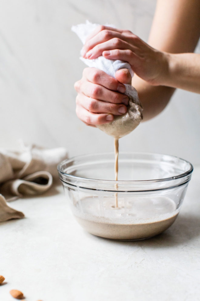 This is a side view of hands squeezing almond milk through a nut milk bag into a glass bowl. The bowl sits on a white counter with a white background. A tan towel is blurred off to the left in the background. A few almonds are scattered on the counter.