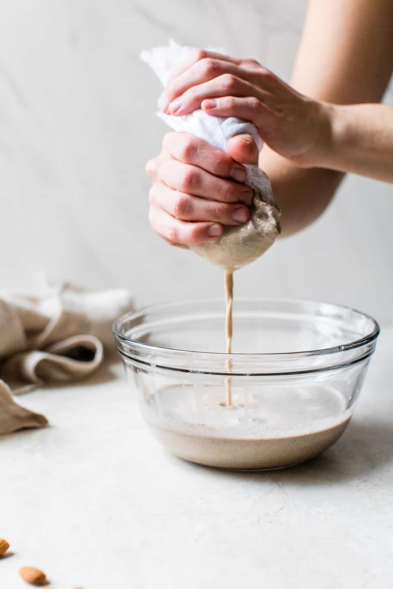 hands squeezing almond milk through a cheese cloth into a bowl on a white counter and white background