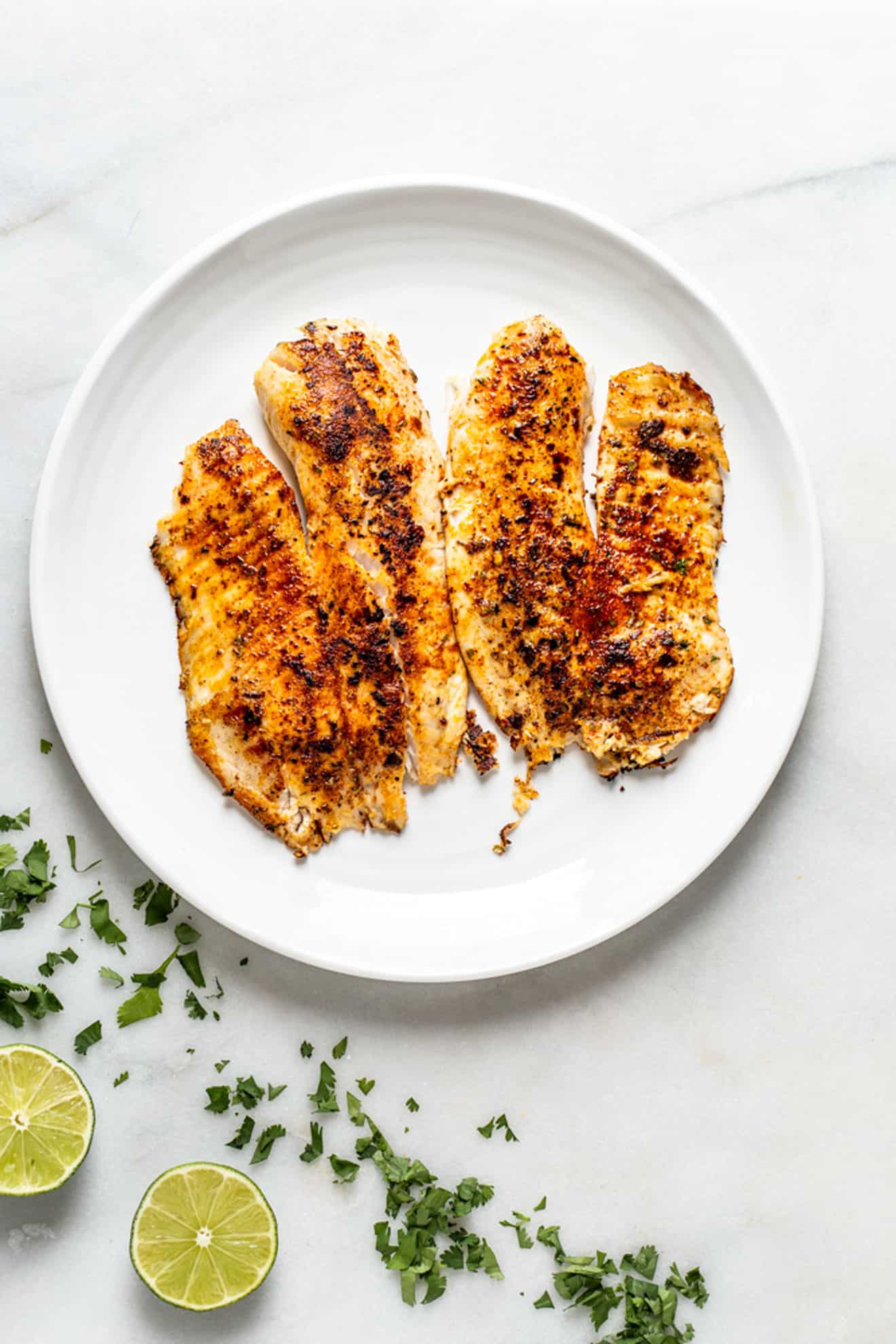 two blackened tilapia filets on a white plate on a marble surface with cilantro and lime wedge
