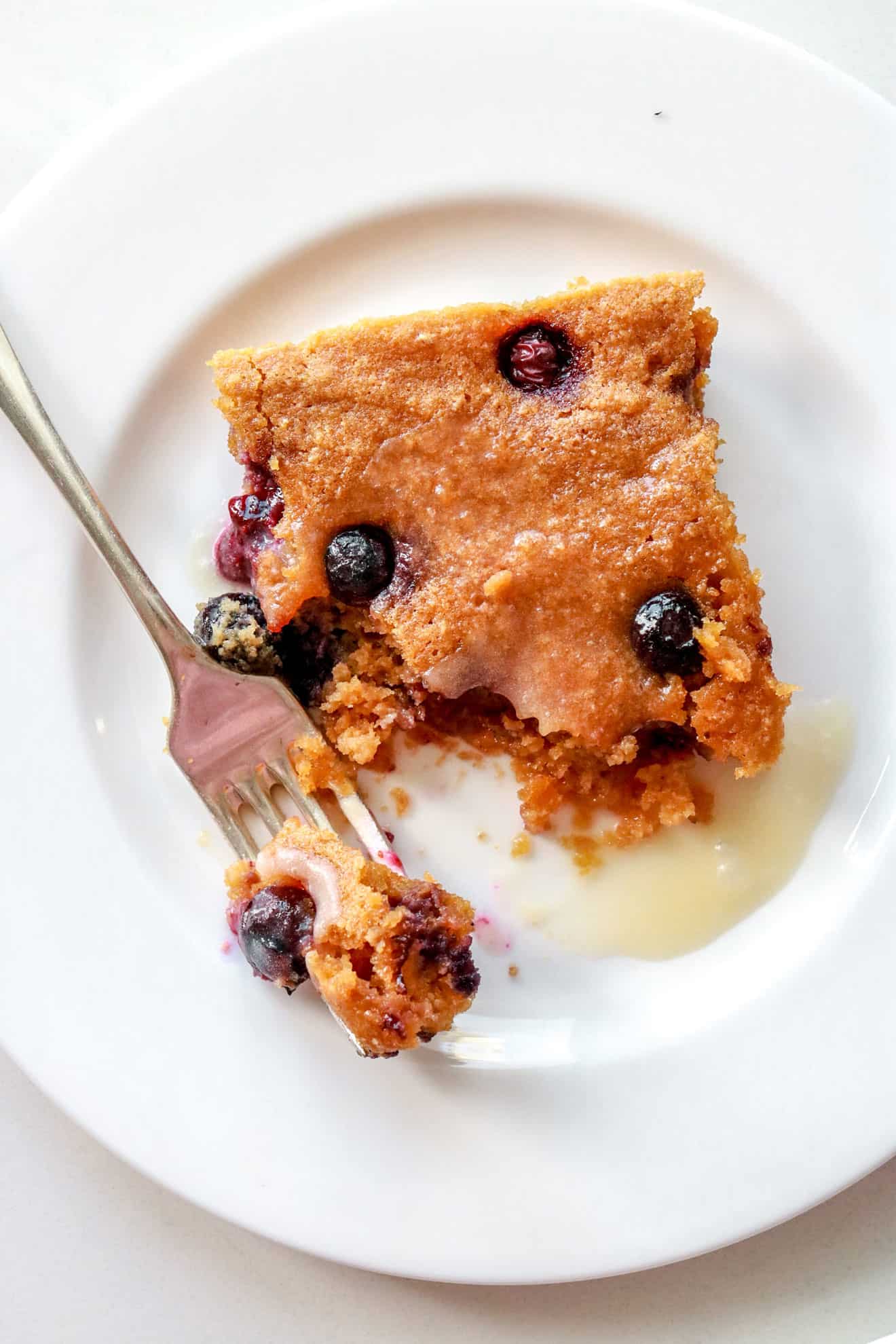 This is an overhead vertical image of a square  cake with blueberries on top sitting on a white plate. The square cake is topped with a white glaze. A fork is coming in from the top corner and angled to lean on the center of the plate with a bite taken out of it. 