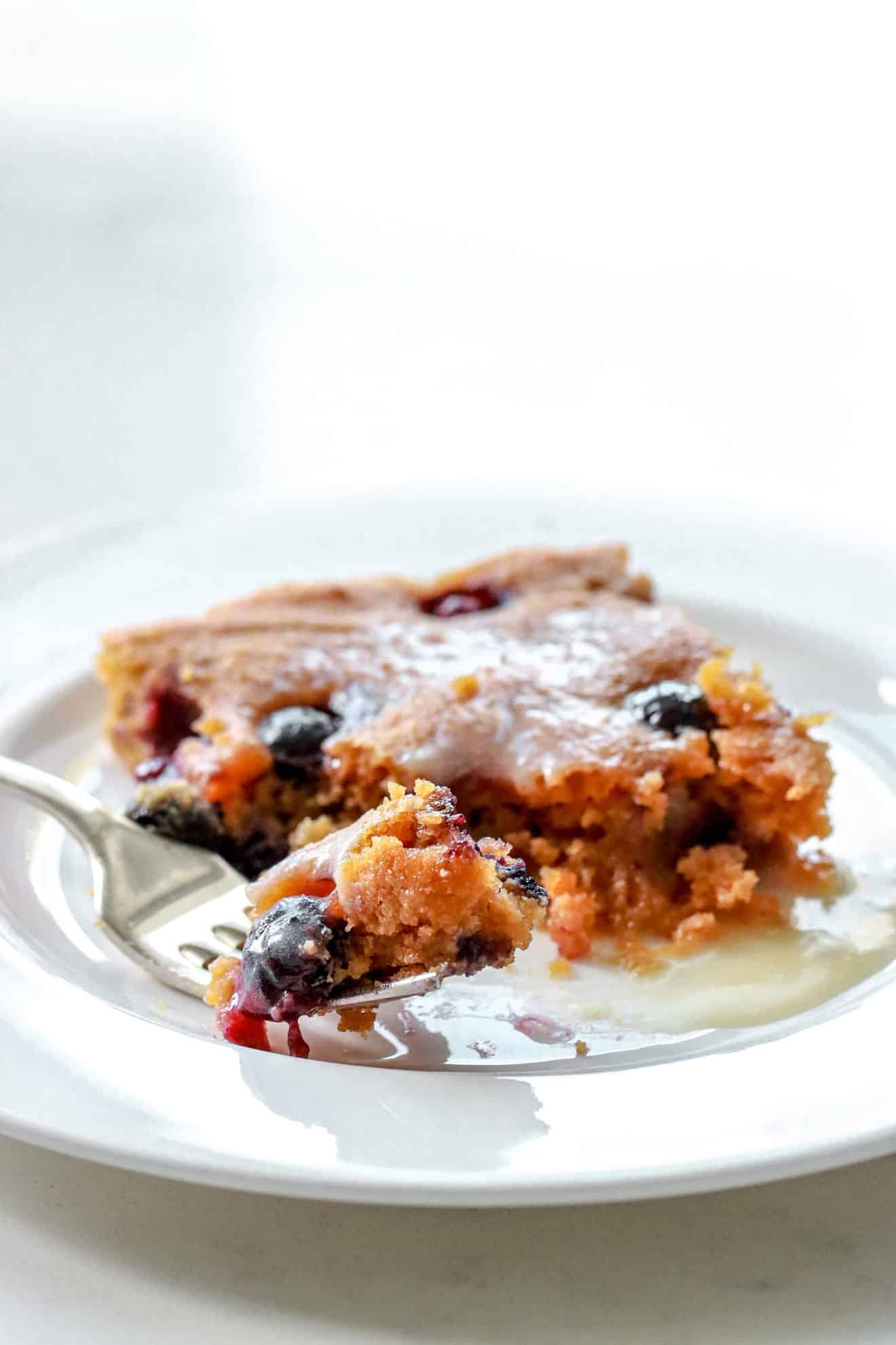 This is a vertical image of a square  cake with blueberries on top sitting on a white plate. The square cake is topped with a white glaze. A fork is coming in from the side and angled to lean on the center of the plate with a bite taken out of it. The image mainly focuses on the bite on the fork. 
