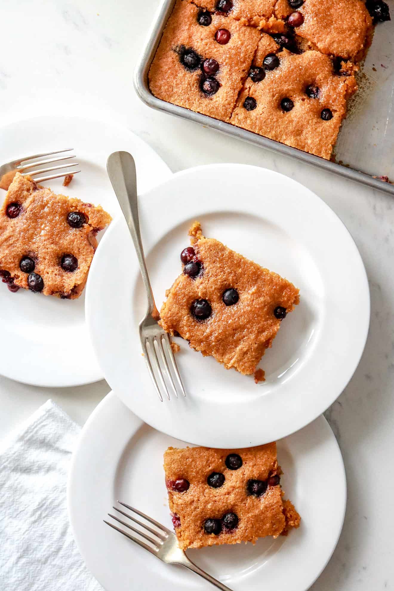 This is an overhead vertical image of three white plates with squares of blueberry cake on them. Each has a silver fork leaning against the plate. To the top right corner is a half sheet pan with more blueberry squares. The sheet pan and plates sit on a white marble surface. 
