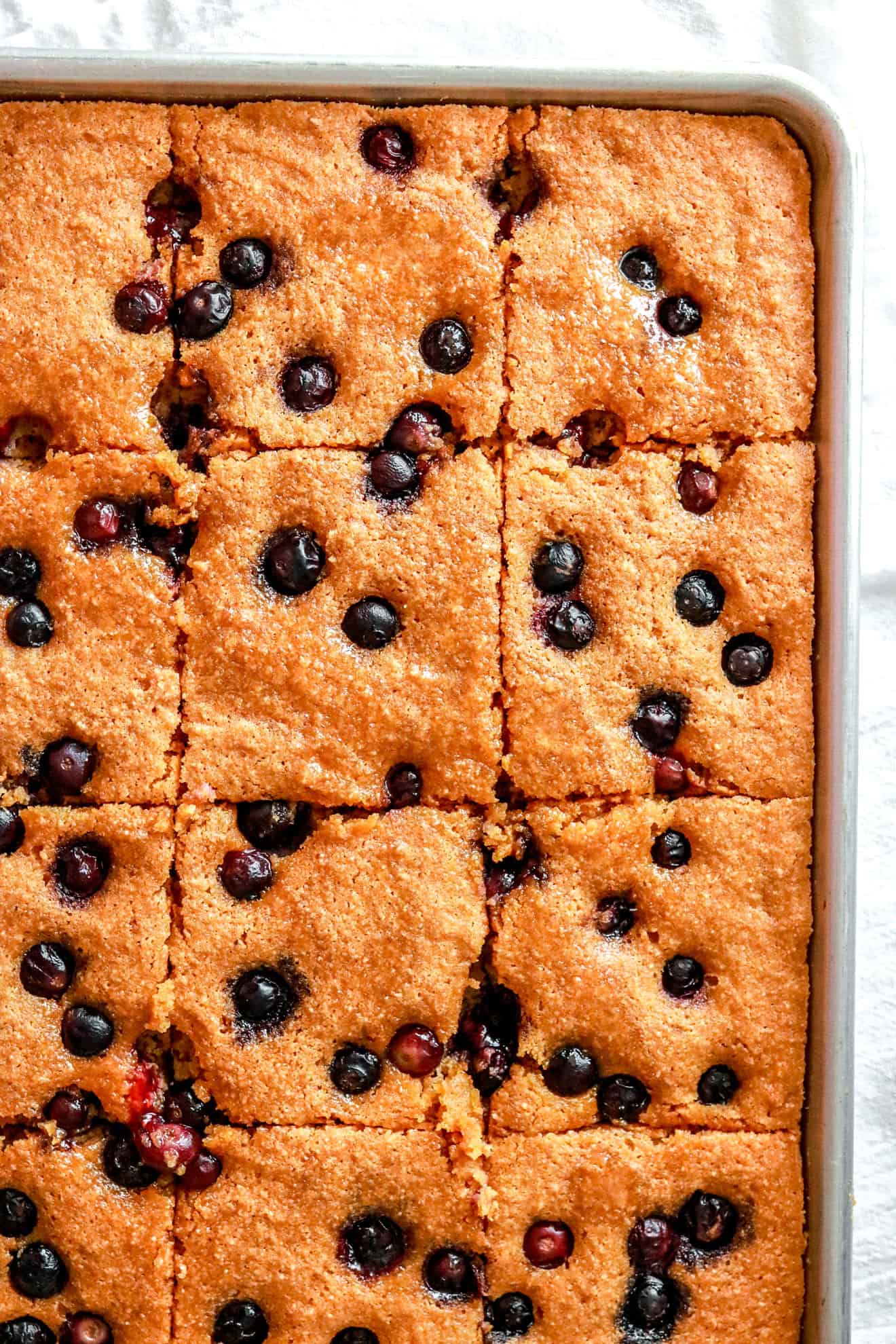 This is an overhead vertical image of a sheet pan with blueberry cake baked. The cake is cut into squares. The sheet pan sits on a light surface. 
