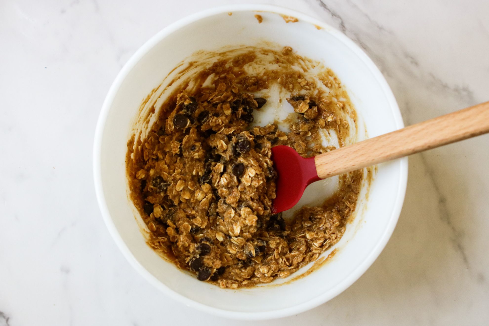This is an overhead horizontal image of a white bowl on a white marble surface. In the bowl is an oatmeal chocolate chip cookie dough mixed together. A red spatula with a wooden handle is inserting into the dough and leaning against the side of the bowl with the whisk pointing to the right side of the image.