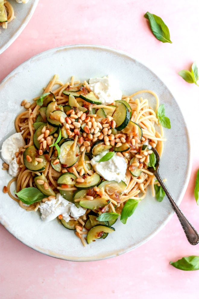 This is an overhead image of a bowl of pasta with zucchini, toasted pine nuts, and burrata. The plate of pasta sits on a pink counter with. fresh basil on top of the pasta and on the counter. 