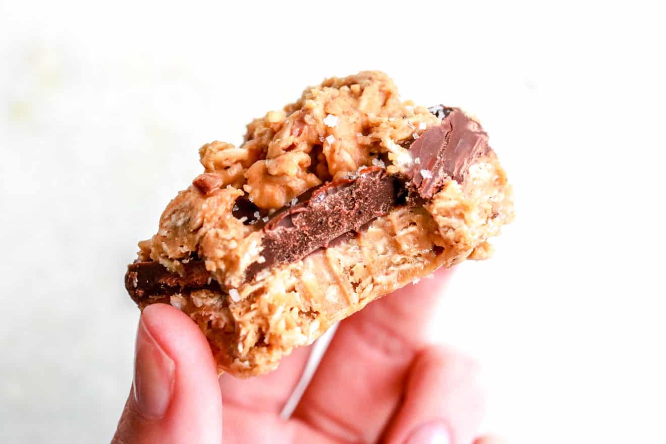A hand is holding an oatmeal bar layered with chocolate, and peanut butter. A bite taken out being held in a hand against a white background.