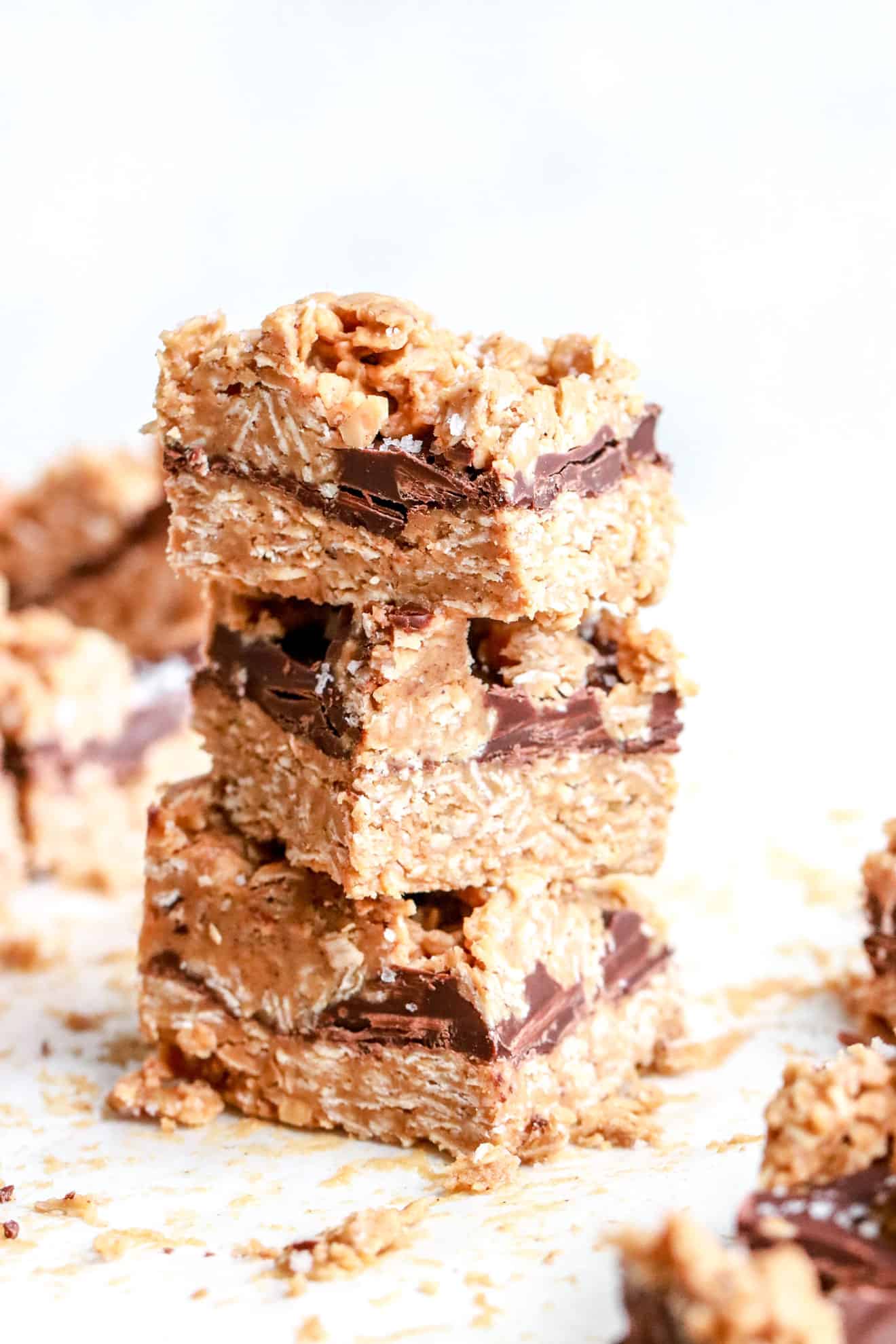 This image is of a stack of no bake oatmeal bars with bars in the foreground and more blurred in the background on white table. The bars have a visible layer of chocolate and are sprinkled with flakey salt.